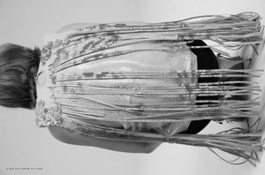 Model seen from behind wearing an elaborate garment with long metallic fringes and sequins against a neutral background.