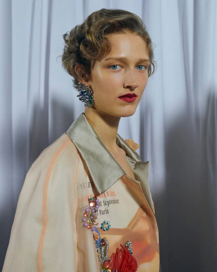 Model with vintage-styled hair wearing embellished jacket and statement earring against white curtain background