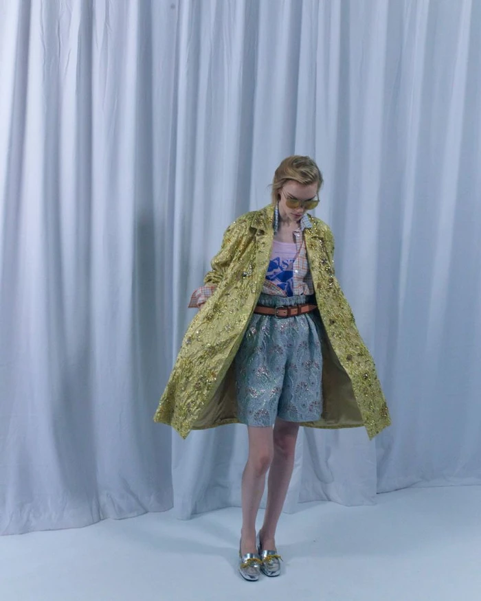 Model in a textured gold coat and patterned shorts standing before white curtains