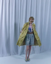 Model in a textured gold coat and patterned shorts standing before white curtains