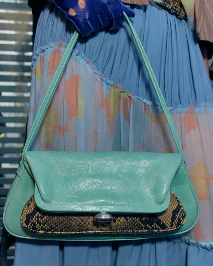 Mint green leather shoulder bag with snakeskin front panel detail held against a blue pleated skirt.