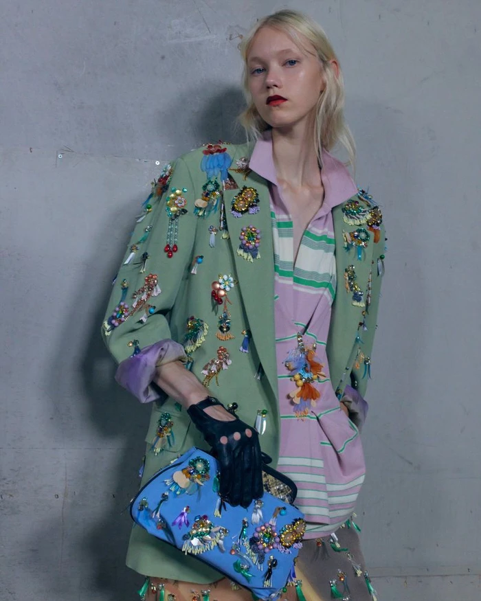 Model in embellished sage green blazer and striped pink top holding a blue handbag against a grey concrete wall