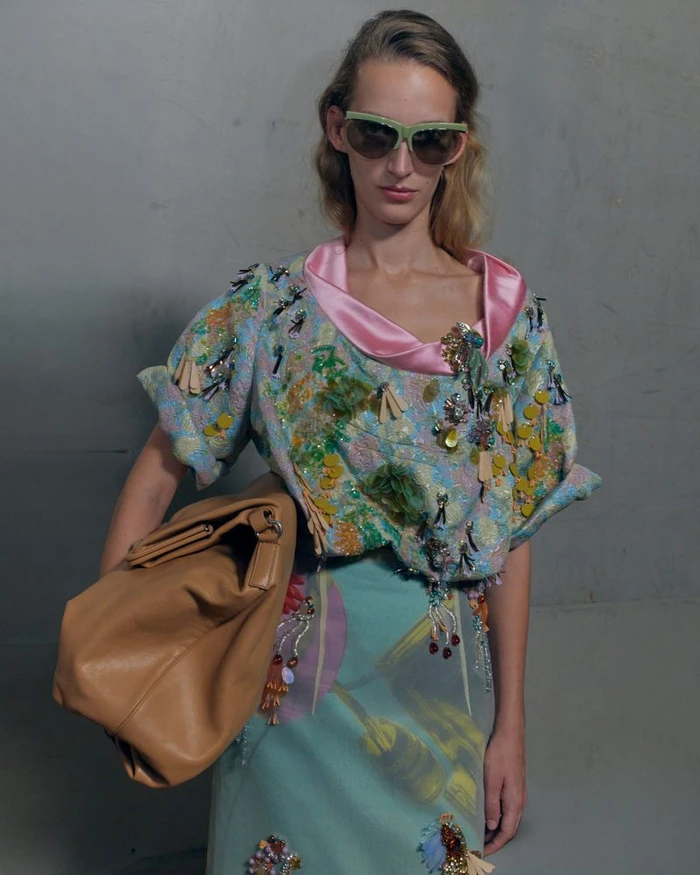 Model wearing embellished patterned top and sunglasses holding a large brown leather bag against a grey background.