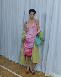 Model in a pink slip dress and olive green coat standing against white curtains for Dries Van Noten SS27