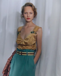 Model in gold satin top with crystal straps and green belted skirt against a white curtain background