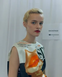 Model with blonde hair and red lipstick wearing a printed graphic top in a backstage setting.