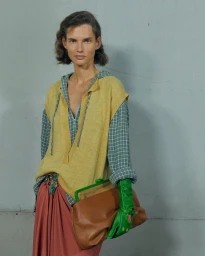 Model in yellow sleeveless knit vest over plaid shirt holding a brown clutch bag against a plain wall