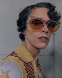 Model wearing amber-tinted sunglasses and a patterned yellow and red knit vest against a plain grey wall.