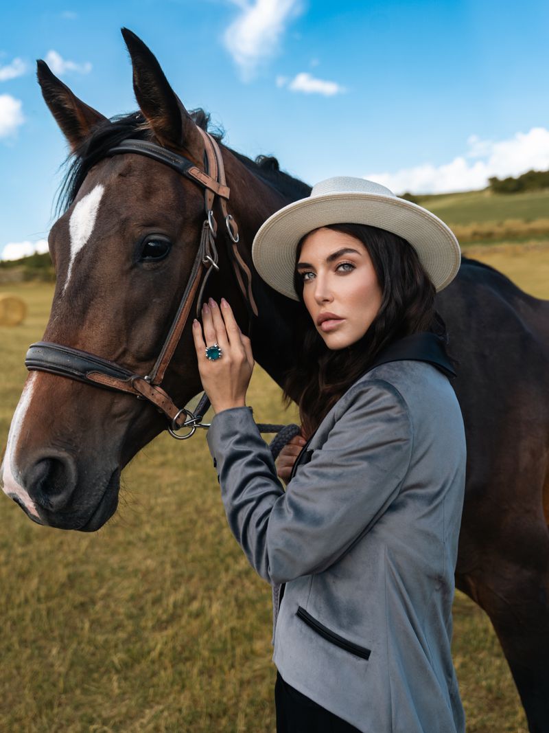 Woman in a grey blazer and hat poses with a dark brown horse in a grassy field under a blue, partly cloudy sky on a sunny day.