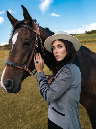 Woman in a grey blazer and hat poses with a dark brown horse in a grassy field under a blue, partly cloudy sky on a sunny day.