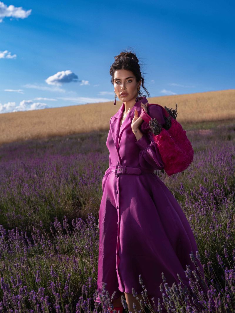 A fashionable woman in a purple coat stands in a lavender field on a sunny day with a bright pink bag slung over her shoulder.