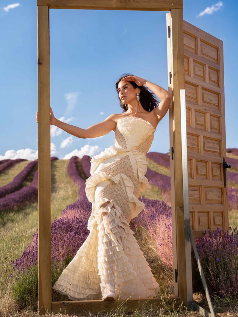 Adult female model posing in ruffled gown in a wooden door frame in a lavender field under a blue sky on a sunny day.