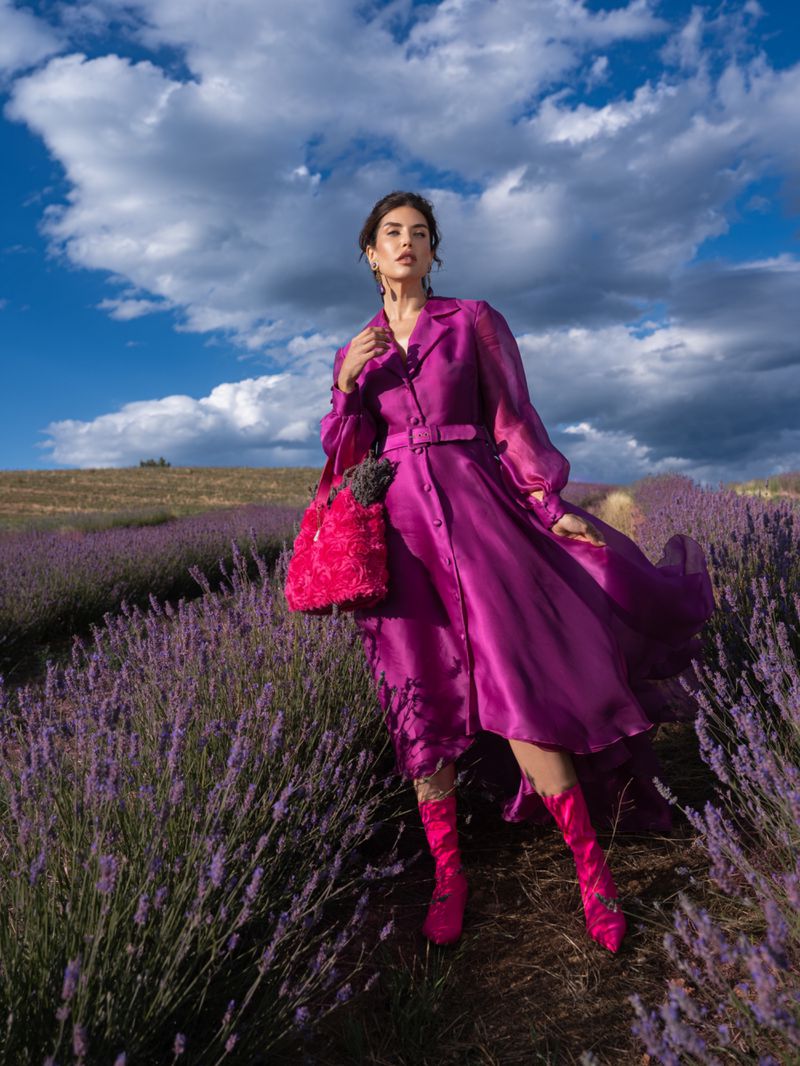Brunette woman in a magenta dress and boots, carrying a flower-filled bag, stands in a lavender field under a partly cloudy sky.