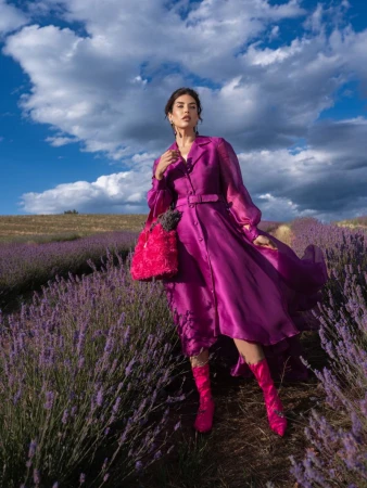 Brunette woman in a magenta dress and boots, carrying a flower-filled bag, stands in a lavender field under a partly cloudy sky.