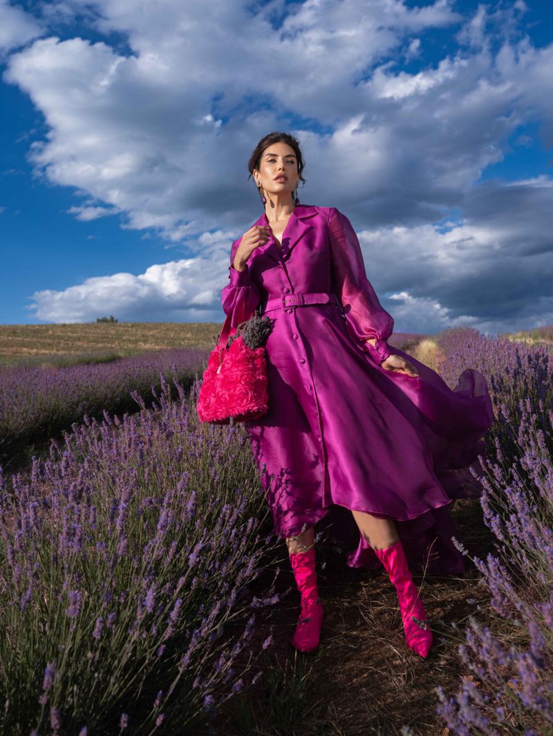 Adult woman in a magenta dress and matching boots stands in a field of lavender under a blue sky with clouds holding a floral bag.