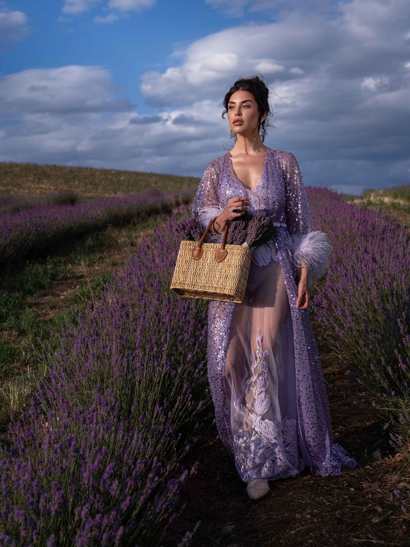 Woman in a purple robe walking through a lavender field, carrying a straw bag, under a cloudy sky, creating a dreamy, natural scene.