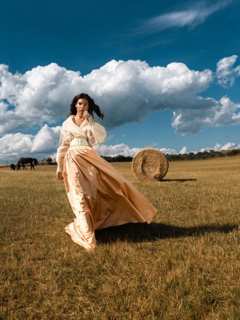 A woman in a gown standing in a grassy field on a sunny day with blue sky, white clouds, and hay bales in a rural environment.