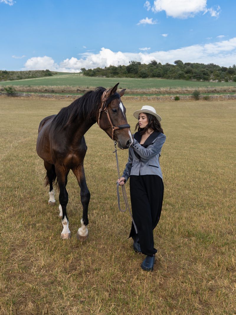 Woman in velvet blazer leads brown horse in sunny field, blue sky with clouds, rural countryside fashion.