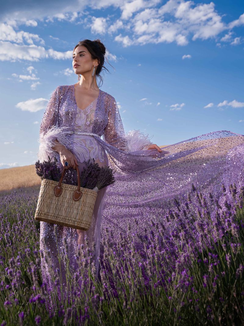 An adult female stands in a lavender field wearing a long, sheer, purple gown and holding a basket of lavender against a partly cloudy blue sky.