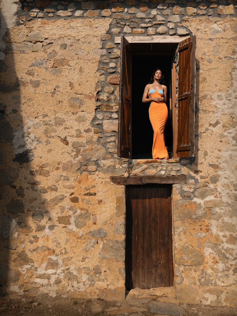 Adult female model in an orange dress stands in a window of a stone building with weathered wooden shutters in natural light.