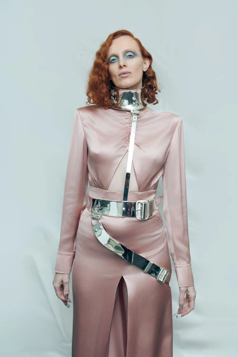 Model with red hair wearing pink satin dress and silver metallic body harness against light backdrop