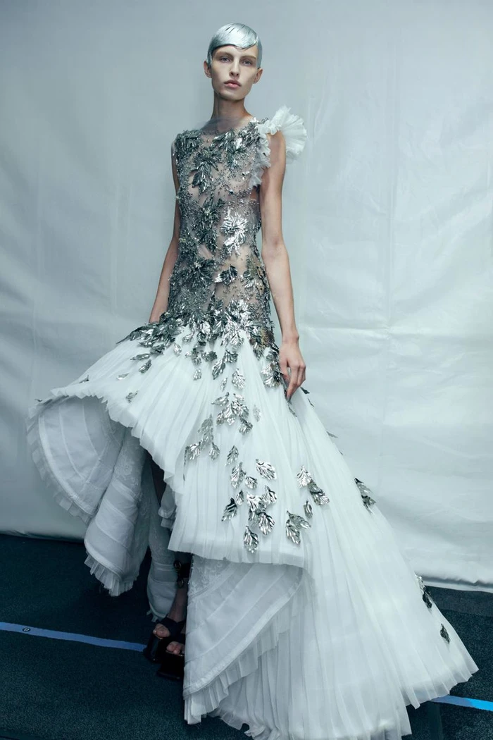 Model in a silver-embellished sheer and white tiered gown with cropped silver hair against a white backdrop.
