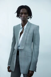 Model with dreadlocks wearing a grey plaid blazer and white shirt against a white seamless background