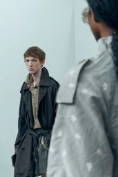 Model wearing dark trench coat standing before a person viewed from behind in a pale studio setting
