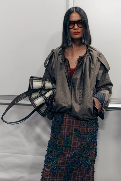 Model wearing large sunglasses and an olive jacket holding a plaid handbag against a plain background.