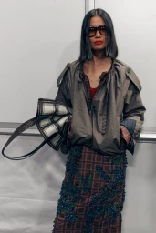 Model wearing large sunglasses and an olive jacket holding a plaid handbag against a plain background.