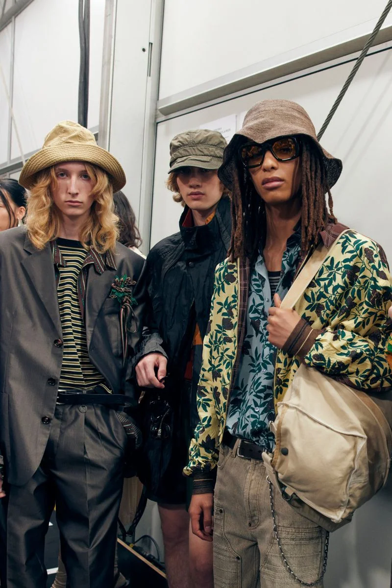 Three models backstage wearing bucket hats, patterned shirts, and layered outerwear during a fashion presentation.