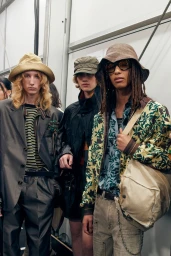 Three models backstage wearing bucket hats, patterned shirts, and layered outerwear during a fashion presentation.