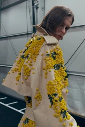 Model wearing a cream trench coat with yellow floral embellishments looking over shoulder