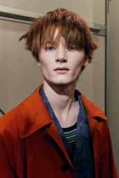 Portrait of a male model with reddish-brown hair wearing an orange coat over a plaid shirt for Burberry SS27