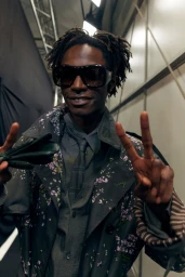 Model wearing a dark floral coat and sunglasses flashing a peace sign backstage for Burberry SS27 BOH