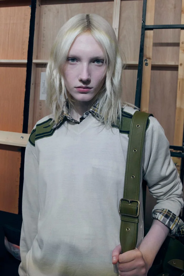 Model with blonde hair wearing cream sweater and plaid shirt backstage at Burberry SS27