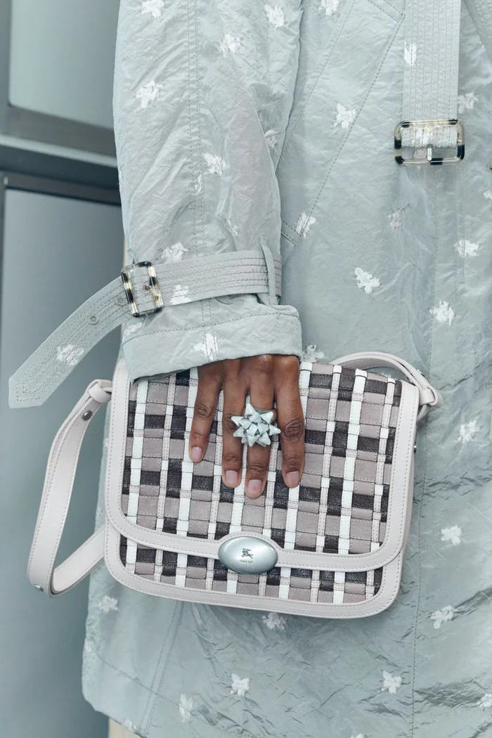 Close-up of a person wearing a pale green patterned coat holding a woven check handbag with a prominent bow ring.