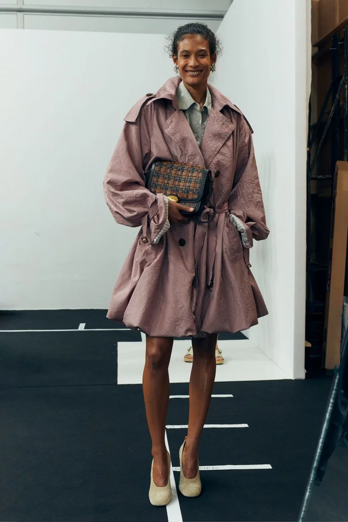 Model wearing a structured dusty rose trench coat holding a patterned handbag in a backstage setting for Burberry SS27