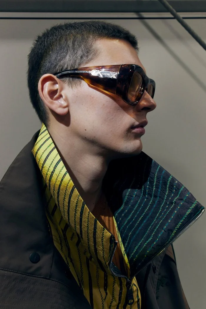 Close-up profile of a male model wearing tortoiseshell sunglasses, a yellow patterned collar, and dark outerwear against a plain wall