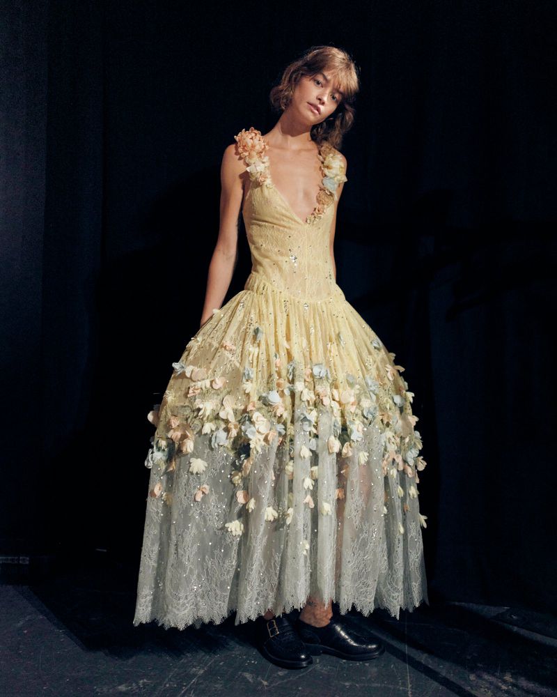 Model in a pale yellow embroidered gown adorned with floral appliqués posing in front of a dark backdrop.