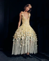 Model in a pale yellow embroidered gown adorned with floral appliqués posing in front of a dark backdrop.