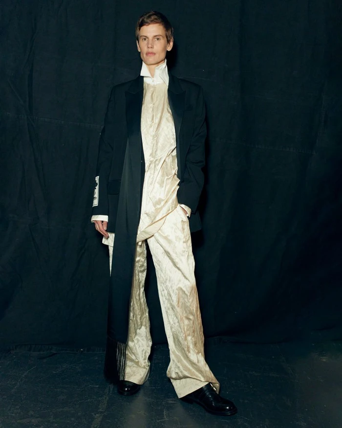Model standing against a dark curtain backdrop wearing a dark blazer over a cream patterned tunic and trousers