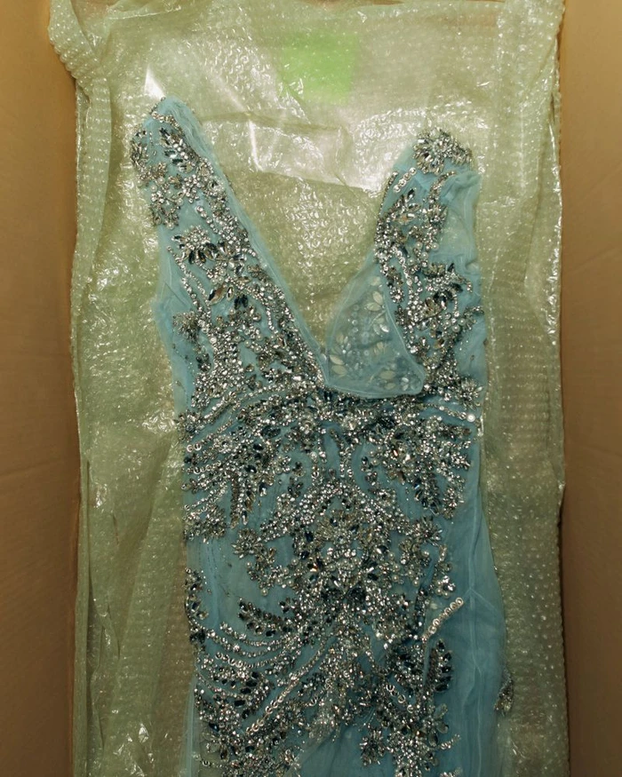 Light blue sleeveless evening dress heavily embellished with silver crystals lying inside a cardboard shipping box lined with bubble wrap.