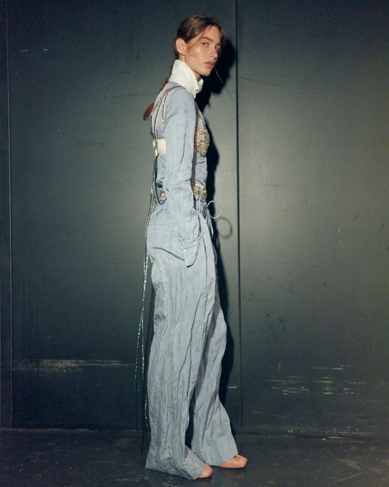 Model standing sideways in a matching light blue textured top and trousers against a dark panel background