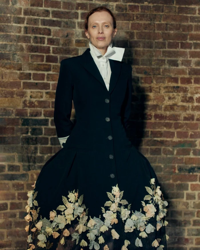Model Karen Elson wearing a black coat with floral embroidery standing against a brick wall for Erdem SS27 BOH