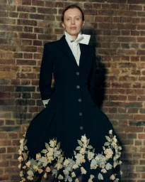 Model Karen Elson wearing a black coat with floral embroidery standing against a brick wall for Erdem SS27 BOH