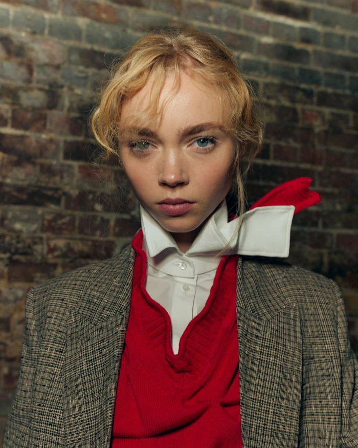 Model with blonde hair wearing a plaid blazer, red sweater, and white collared shirt against a brick wall background