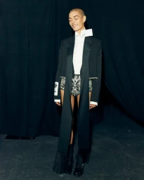 Model with a shaved head wearing a cropped black jacket and white button-down shirt against a dark fabric backdrop.