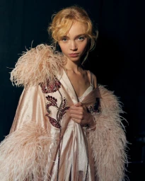 Model wearing a pink feather-trimmed robe with beaded floral embellishments against a dark background