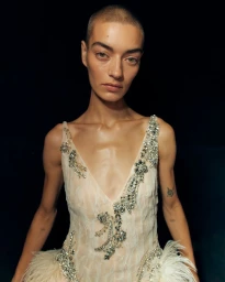 Model with a buzzcut wearing an embellished cream dress with crystal detailing and feather trim against a dark background